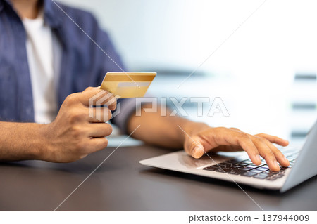 Close-up photo of the hands of a young man sitting at a desk in an office, holding a gold credit card and using a laptop. Close-up photo of the hands of a young man sitting at a desk in an office, holding a gold credit card and using a laptop. 137944009