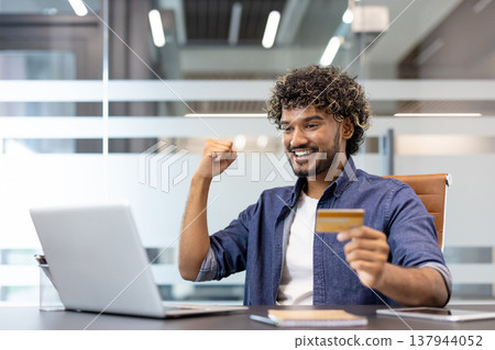 Happy young Indian man sitting at a desk in the office, looking at the laptop screen, holding a credit card in his hand and rejoicing in his success. Happy young Indian man sitting at a desk in the office, looking at the laptop screen, holding a credit card in his hand and rejoicing in his success. 137944052