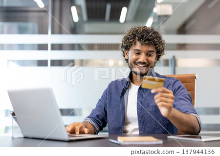 A young Indian man is sitting at a desk in an office, working on a laptop and looking at a credit card he is holding in his hand. A young Indian man is sitting at a desk in an office, working on a laptop and looking at a credit card he is holding in his hand. 137944136