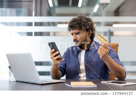Worried young Indian man sitting at a desk in the office with a credit card in his hand and looking at his phone in frustration. 137944137
