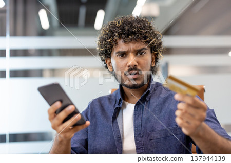 Close-up portrait of a young Indian man in a denim shirt sitting in an office, holding a credit card and phone in his hand, looking worriedly at the camera. Close-up portrait of a young Indian man in a denim shirt sitting in an office, holding a credit card and phone in his hand, looking worriedly at the camera. 137944139