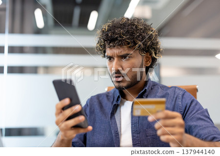 Close-up photo of a worried young Indian man in the office, holding a credit card and looking at his phone screen in frustration. Close-up photo of a worried young Indian man in the office, holding a credit card and looking at his phone screen in frustration. 137944140