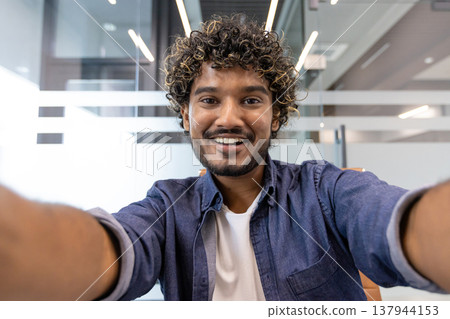 Close-up portrait of a young Indian man in an office, smiling and talking to the camera he is holding. 137944153