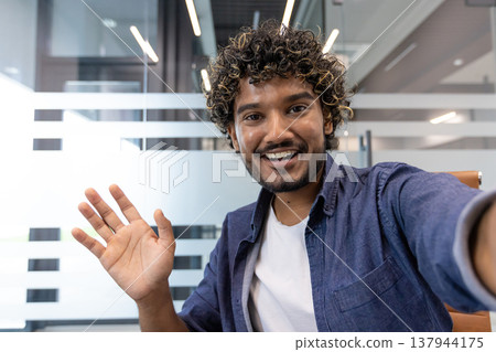 Close-up portrait of a young Indian man in an office, talking and greeting the camera he is holding in his hand. Close-up portrait of a young Indian man in an office, talking and greeting the camera he is holding in his hand. 137944175