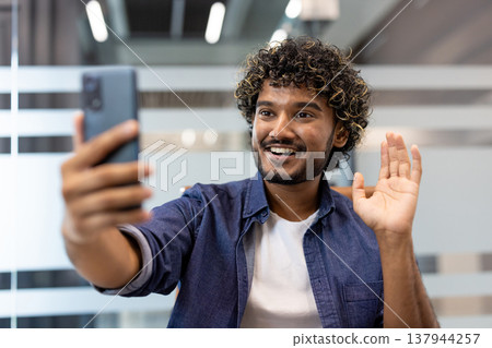Close-up photo of a young Indian man sitting in an office at his desk, smiling and talking on the camera phone he is holding in his hand. Close-up photo of a young Indian man sitting in an office at his desk, smiling and talking on the camera phone he is holding in his hand. 137944257