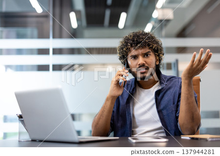 Close-up portrait of worried Indian young man sitting in office and talking on mobile phone, spreading his hands in frustration at camera. Close-up portrait of worried Indian young man sitting in office and talking on mobile phone, spreading his hands in frustration at camera. 137944281