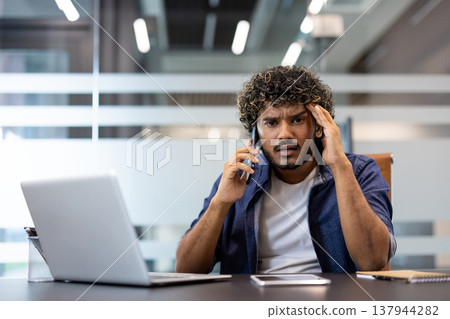 Portrait of a worried young Indian man sitting at an office desk, talking on the phone and looking at the camera in frustration, holding his head. Portrait of a worried young Indian man sitting at an office desk, talking on the phone and looking at the camera in frustration, holding his head. 137944282
