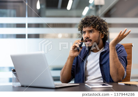 Angry and frustrated young Indian man sitting at a desk in the office and talking emotionally on a mobile phone. Angry and frustrated young Indian man sitting at a desk in the office and talking emotionally on a mobile phone. 137944286