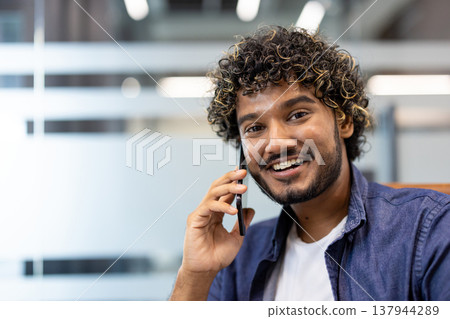Close-up photo of a young Indian man sitting in an office and smiling while talking on the phone. Close-up photo of a young Indian man sitting in an office and smiling while talking on the phone. 137944289