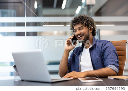 A young Indian man is sitting in an office at a desk in front of a laptop and talking on the phone with a smile. A young Indian man is sitting in an office at a desk in front of a laptop and talking on the phone with a smile. 137944293