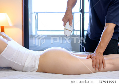 A woman gets help with VIO hair removal from her partner (boyfriend, husband) while lying face down, using a home hair removal device, from a side view, with a strobe light. A woman gets help with VIO hair removal from her partner (boyfriend, husband) while lying face down, using a home hair removal device, from a side view, with a strobe light. 137944994