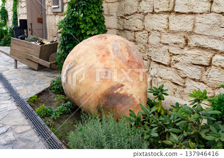 A large, brown, ceramic vase is sitting on the ground next to some plants A large, brown, ceramic vase is sitting on the ground next to some plants 137946416