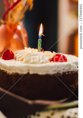 Chocolate birthday cake with whipped cream, cherries, and a burning candle on a table. Holiday, anniversary, and celebration designs. Chocolate birthday cake with whipped cream, cherries, and a burning candle on a table. Holiday, anniversary, and celebration designs. 137946992