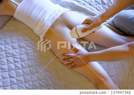 Women getting help with VIO hair removal from their partners (boyfriends, husbands) while lying face down using a home hair removal device. 137947368