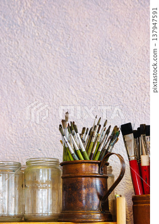 Closeup of assorted paintbrushes stored in a vintage copper pitcher and empty glass jars against a textured wall Closeup of assorted paintbrushes stored in a vintage copper pitcher and empty glass jars against a textured wall 137947591