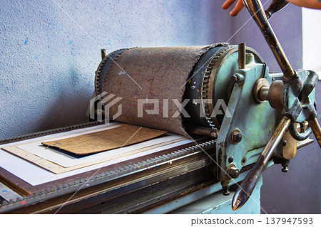 Closeup of a hand operated manual printing press with a layered printmaking plate and paper running through the rollers. 137947593