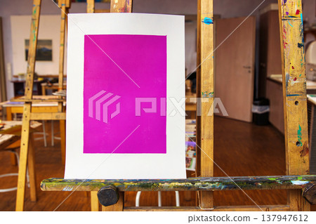 A single magenta monoprint is centered on a white sheet of paper, standing upright on a well used wooden easel in an art classroom. A single magenta monoprint is centered on a white sheet of paper, standing upright on a well used wooden easel in an art classroom. 137947612