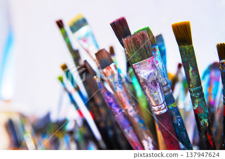 A cluster of well used paintbrushes with colorful dried paint on their handles and bristles in container in creative art studio. 137947624