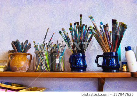 Various paintbrushes of different shapes and size, arranged in ceramic mugs and glass jars on a wooden shelf against a textured wall. Various paintbrushes of different shapes and size, arranged in ceramic mugs and glass jars on a wooden shelf against a textured wall. 137947625