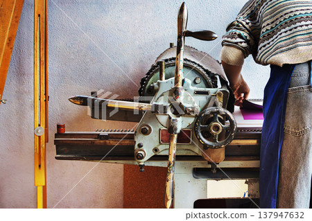 A printmaker in a patterned sweater and apron operates a traditional mechanical printing press. Back view of the artist during the printmaking process 137947632