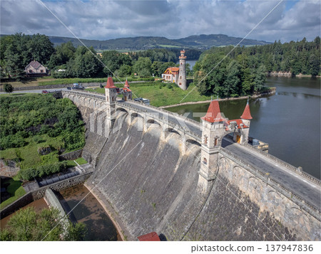 Aerial view of Les Kralovstvi dam with stone towers and red roofs spanning a river reservoir, surrounded by forested hills and countryside under dramatic cloudy sky in Czech Republic. 137947836