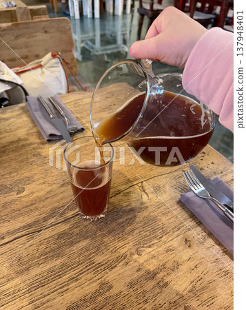 Hand pours kvass from glass jug into tall glass on wooden table in restaurant setting, traditional fermented drink with dark color and foam. Beverage serving, drink presentation, restaurant menu Hand pours kvass from glass jug into tall glass on wooden table in restaurant setting, traditional fermented drink with dark color and foam. Beverage serving, drink presentation, restaurant menu 137948401
