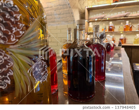 Glass bottles with infused drinks in amber and red tones stand on bar counter in rustic interior with shelves and warm lighting, beverage assortment with rich colors. Beverage assortment, product Glass bottles with infused drinks in amber and red tones stand on bar counter in rustic interior with shelves and warm lighting, beverage assortment with rich colors. Beverage assortment, product 137948405