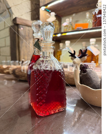 Glass bottle with red infused drink stands on wooden counter with other bottles, decor in rustic interior, beverage presentation with rich color and texture. Beverage styling, product, bar menu Glass bottle with red infused drink stands on wooden counter with other bottles, decor in rustic interior, beverage presentation with rich color and texture. Beverage styling, product, bar menu 137948422
