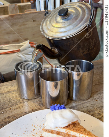 Hot drink poured from metal kettle into cup on wooden table with metal mugs and dessert in restaurant setting under warm light. Beverage preparation, tea service, food styling, restaurant marketing Hot drink poured from metal kettle into cup on wooden table with metal mugs and dessert in restaurant setting under warm light. Beverage preparation, tea service, food styling, restaurant marketing 137948433