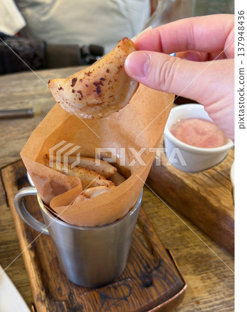 Hand holds small fried dumpling above metal cup with paper liner and sauce on wooden table in restaurant setting, crispy snack with golden texture. Food serving, street food menu, snack 137948436