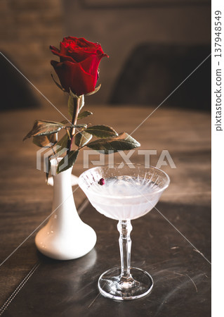Top view of a crystal cocktail glass and a red rose in a white vase on a dark wooden table. Moody still life for romantic and celebration designs. Top view of a crystal cocktail glass and a red rose in a white vase on a dark wooden table. Moody still life for romantic and celebration designs. 137948549