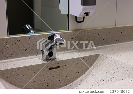 Modern sensor faucet mounted on a gray stone basin, mirror, and soap dispenser in public restroom. Modern sensor faucet mounted on a gray stone basin, mirror, and soap dispenser in public restroom. 137948622