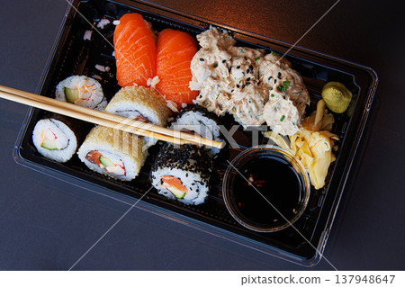 A takeaway sushi meal in a black plastic tray with salmon nigiri, maki rolls, ginger, wasabi, and soy sauce with chopsticks over the tray. Selective focus 137948647