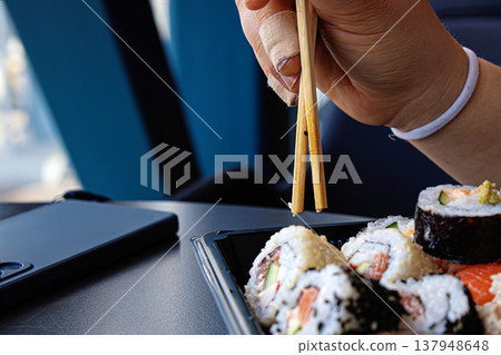 Hand with plaster using chopsticks to eat sushi from takeout container on dark table. Casual dining scene with a smartphone nearby. Selective focus 137948648