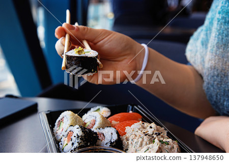 Closeup of the person hand picking up a sushi roll with chopsticks over a black tray in a modern cafe. Selective focus 137948650