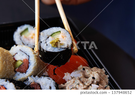 Chopsticks grasping sushi roll with cucumber, avocade and salmon. Maki  being picked up with chopsticks.  Selective focus 137948652