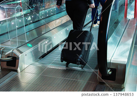 Traveler dragging suitcase on airport walkway. Passenger walks across a moving floor in a port terminal pulling a rolling luggage. Traveler dragging suitcase on airport walkway. Passenger walks across a moving floor in a port terminal pulling a rolling luggage. 137948653