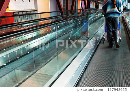 Traveler pushing a luggage trolley on an airport walkway. Passenger walks with travel cart across a moving floor in a modern airport terminal Traveler pushing a luggage trolley on an airport walkway. Passenger walks with travel cart across a moving floor in a modern airport terminal 137948655