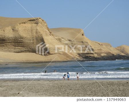 Sandy beach with people playing in ocean waves beneath dramatic desert cliffs. Sandy beach with people playing in ocean waves beneath dramatic desert cliffs. 137949063