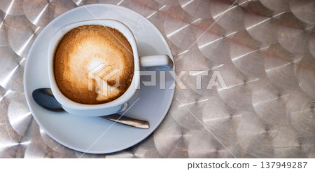 Top view of a cappuccino with creamy milk foam in a square white ceramic cup on a saucer served on a metallic table in outdoor cafe. Selective focus. Top view of a cappuccino with creamy milk foam in a square white ceramic cup on a saucer served on a metallic table in outdoor cafe. Selective focus. 137949287