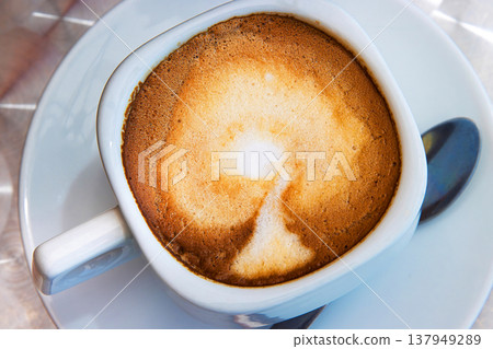 Closeup view of a cappuccino with creamy milk foam in a square white ceramic cup, placed on a saucer. Overhead view. Selective focus Closeup view of a cappuccino with creamy milk foam in a square white ceramic cup, placed on a saucer. Overhead view. Selective focus 137949289