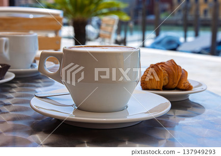 Two white ceramic cups of cappuccino with creamy foam served with italian pastries on a metal table at an outdoor cafe in Murano, Italy on a hot sunny summer day 137949293