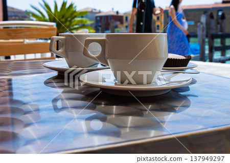 Two white ceramic cups of cappuccino served with italian pastries on a metal table at an outdoor cafe in Murano, Italy on a hot sunny summer day. Two white ceramic cups of cappuccino served with italian pastries on a metal table at an outdoor cafe in Murano, Italy on a hot sunny summer day. 137949297