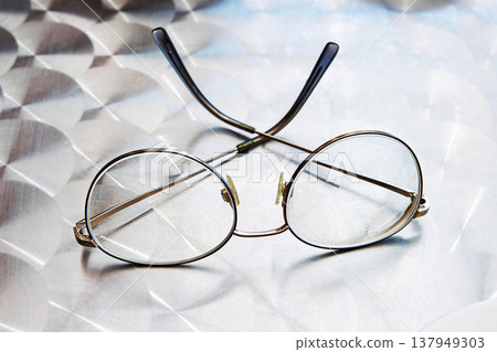 A pair of vintage style round eyeglasses with thin metal frames lies on a geometric metallic surface with a reflective pattern. Selective focus A pair of vintage style round eyeglasses with thin metal frames lies on a geometric metallic surface with a reflective pattern. Selective focus 137949303