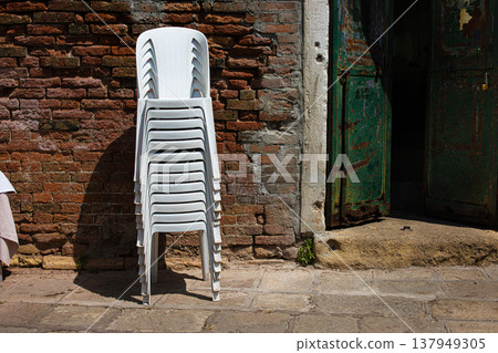Stacked white plastic chairs for restaurant terrace by rustic brick wall in Murano Italy. Stacked white plastic chairs for restaurant terrace by rustic brick wall in Murano Italy. 137949305