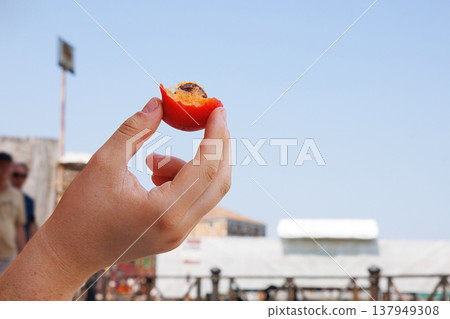 Closeup of a young woman hand holding a partially eaten ripe orange apricot with stone against an old Italian town with clear blue sky on blurred background Closeup of a young woman hand holding a partially eaten ripe orange apricot with stone against an old Italian town with clear blue sky on blurred background 137949308