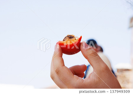 Closeup of a young woman hand holding a partially eaten ripe orange apricot with stone  against clear sky and people on blurred background. Selective focus 137949309