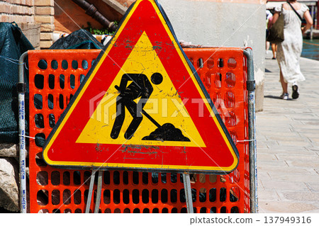 Murano, Province of Venice, Italy, June 14th, 2025. Closeup of a road work warning sign in the construction zone on a city sidewalk with an orange safety barrier and blurred pedestrian Murano, Province of Venice, Italy, June 14th, 2025. Closeup of a road work warning sign in the construction zone on a city sidewalk with an orange safety barrier and blurred pedestrian 137949316