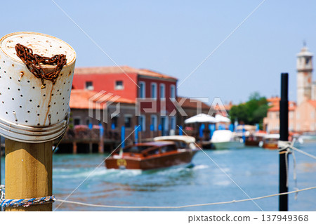Murano, Province of Venice, Italy, June 14th, 2025. Mooring post with plastic fishing bucket  with rusty chain and a motorboat moving along the turquoise canal water passing  colorful buildings. 137949368