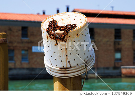A reused plastic bucket with holes, transformed into a fishing container, hangs on an old wooden mooring post known as a palina Canal in Murano, Venetian Lagoon A reused plastic bucket with holes, transformed into a fishing container, hangs on an old wooden mooring post known as a palina Canal in Murano, Venetian Lagoon 137949369